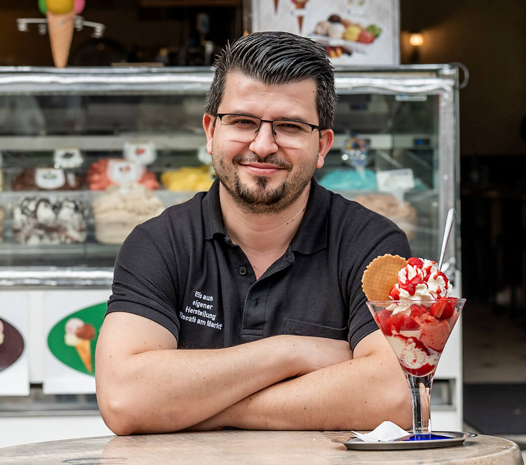 Tiim Gafur, Inhaber des Eiscafés am Markt in Herzogenaurach, sitzt am Tisch seines Cafés im Freien und hat einen Eisbecher vor sich stehen.