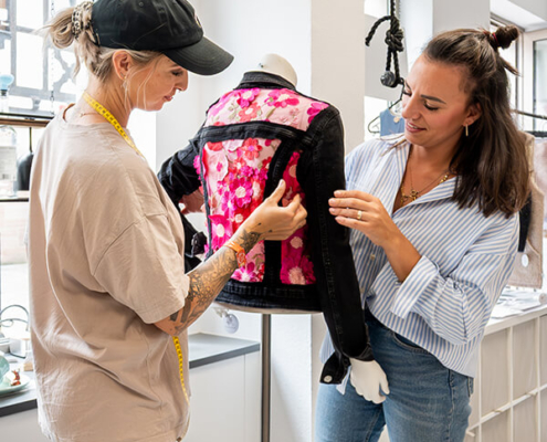 Laura Langer und Miri Zeitschel in der Wunderschmiede, sie richten ein exklusive selbst geschneiderte Jeansjacke an einer Kleiderpuppe im Schaufenster her